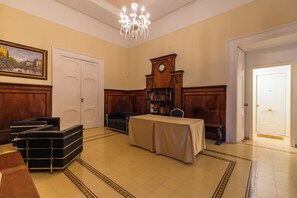 A room with a table covered by a tablecloth, a bookshelf, a clock, and a chandelier.