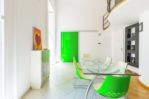 Uma área de jantar moderna com mesa de vidro e cadeiras verdes, um armário branco com pintura de coração, e uma porta verde ao fundo.