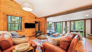 A cozy log cabin interior with a fireplace, leather sofas, and large windows offering a forest view. - Perfect for couples! Modern, Private + Hot Tub, Fire pit, Fireplace & King Bed (Maggie Valley)
