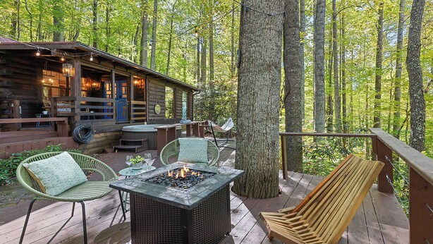 A wooden deck with outdoor seating, a fire pit, and a log cabin in a forested area. - Perfect for couples! Modern, Private + Hot Tub, Fire pit, Fireplace & King Bed (Maggie Valley)