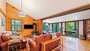 A cozy cabin interior with a fireplace, leather sofa, and large windows offering a view of the forest. - Perfect for couples! Modern, Private + Hot Tub, Fire pit, Fireplace & King Bed (Maggie Valley)