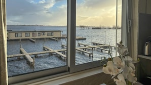 A view through a window of a building with a dock extending into a body of water, surrounded by trees and a cloudy sky. - Visserslust Rooms, Appartments and Houseboats (Vinkeveen)
