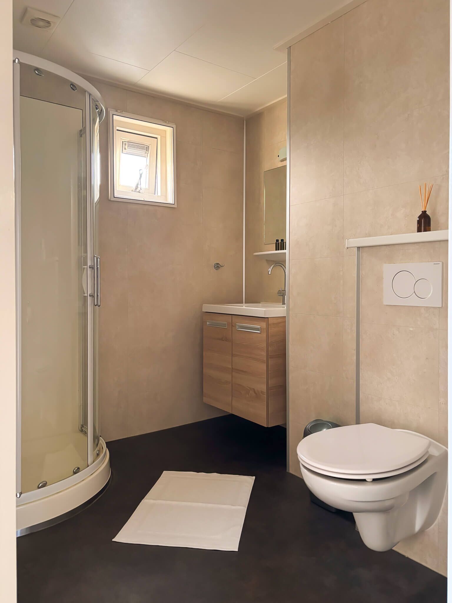 A modern bathroom with a glass shower enclosure, a wooden vanity, a toilet, and a small window providing natural light.