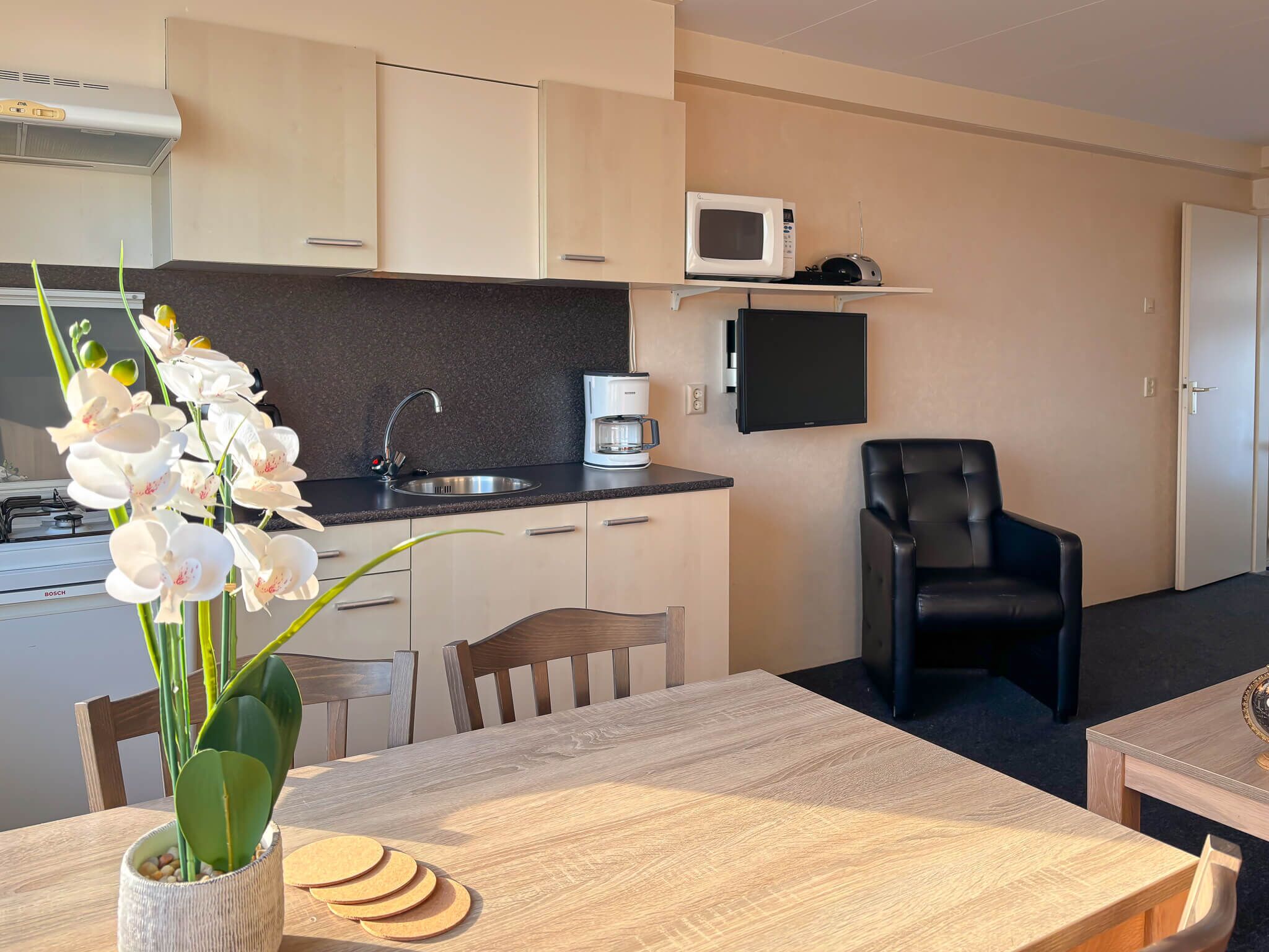 A modern kitchen with a microwave, coffee maker, and a flat-screen TV. There is a dining table with a vase of white flowers and a chair.
