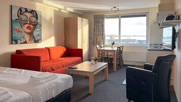 A hotel room with a bed, a red sofa, a dining table with chairs, a black armchair, a painting on the wall, and a view of the city through the window.