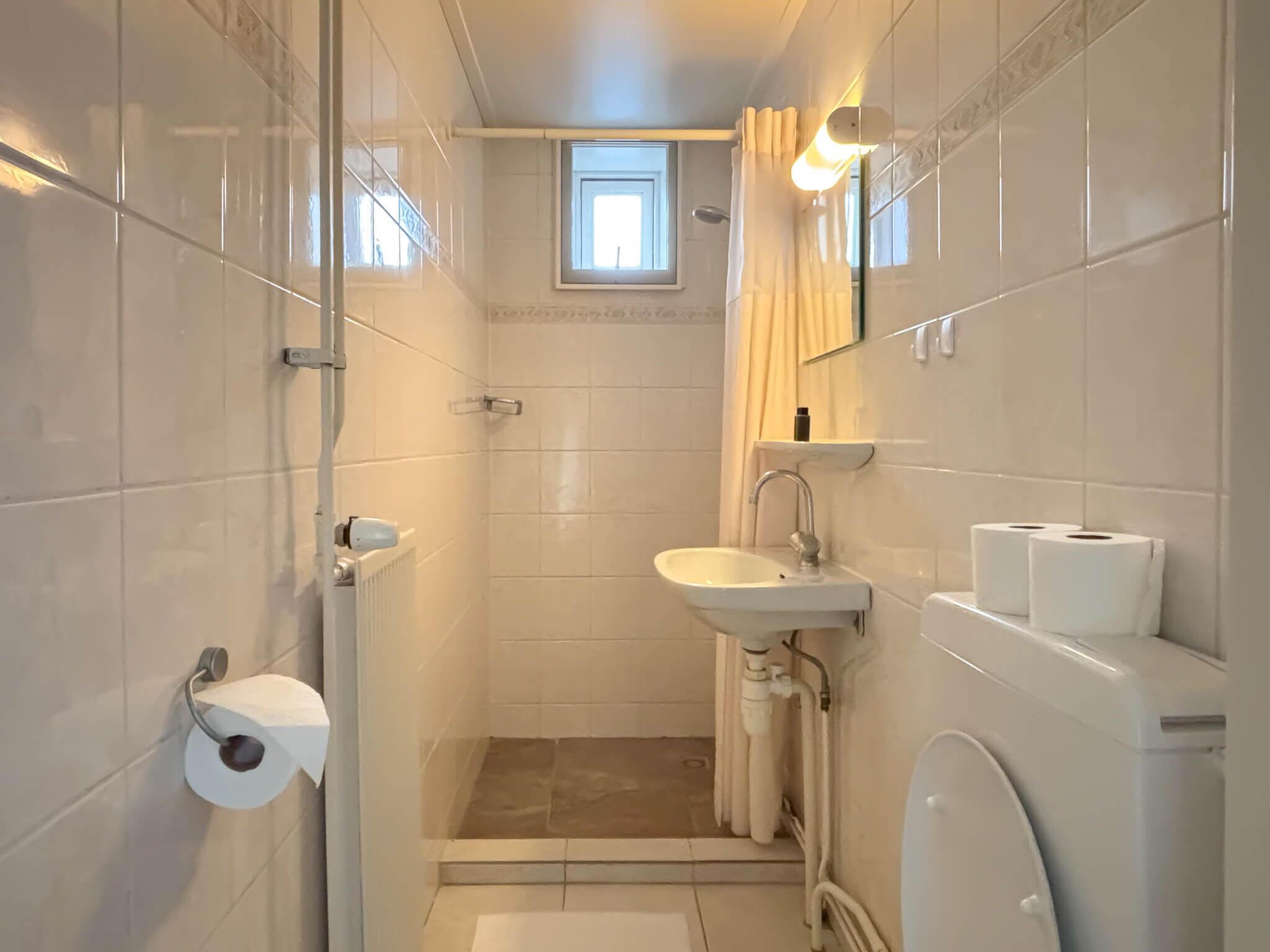 A small, narrow bathroom with a toilet, sink, and shower. There is a window above the shower, and a towel rack is visible on the wall.