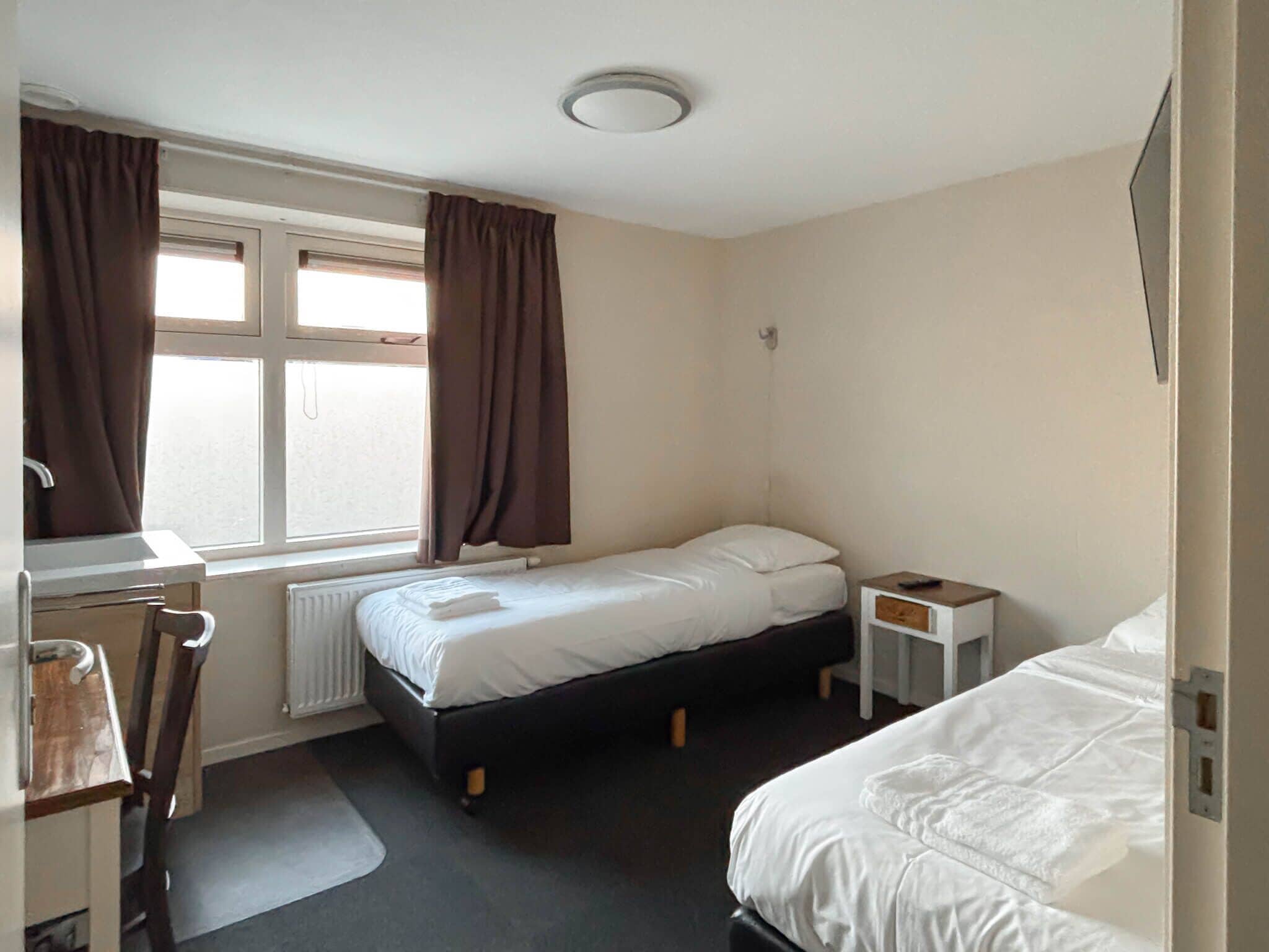 A small, clean room with two beds, a desk, and a window with curtains.
