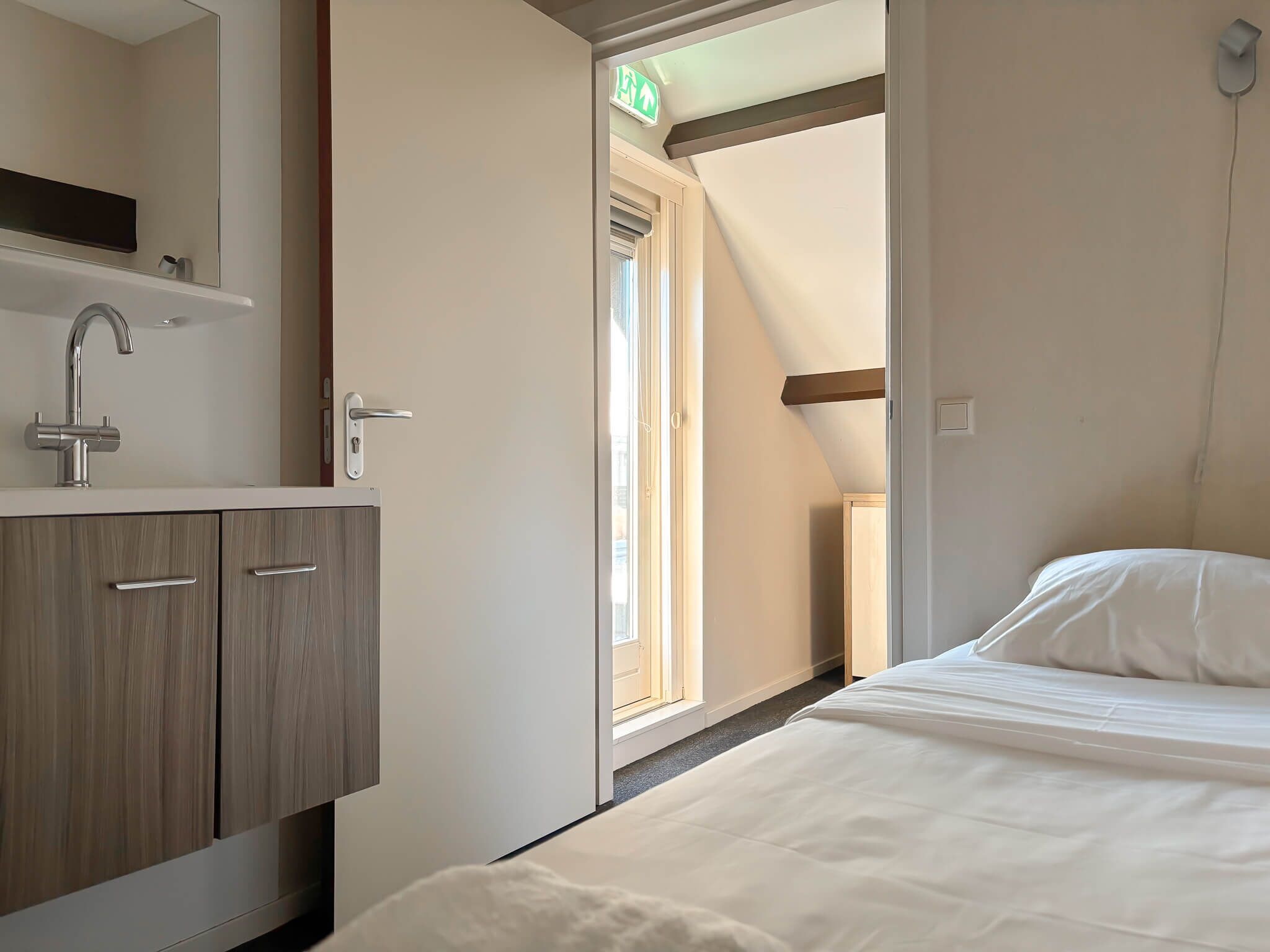 A hotel room with a bed, a bathroom with a sink and cabinets, and a window with an exit sign.