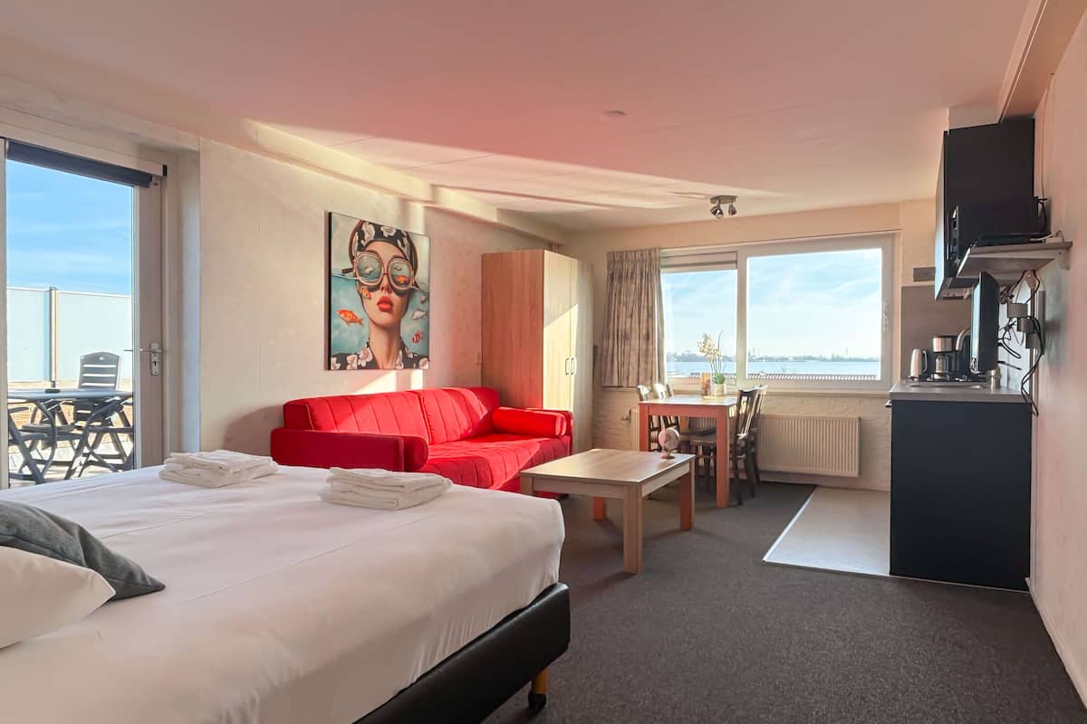 A hotel room with a bed, a red sofa, a dining table, and a view of the sea.