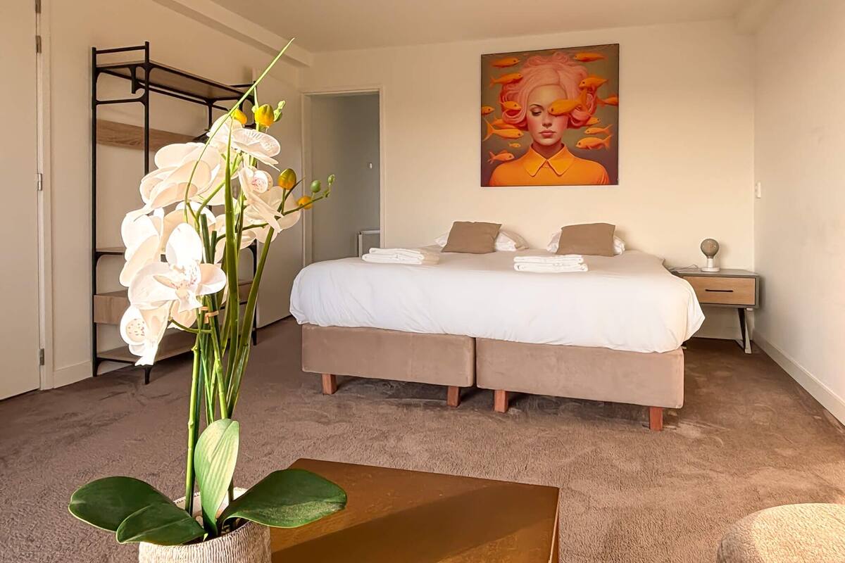 A bedroom with a bed, a nightstand, a lamp, a painting, and a potted orchid.