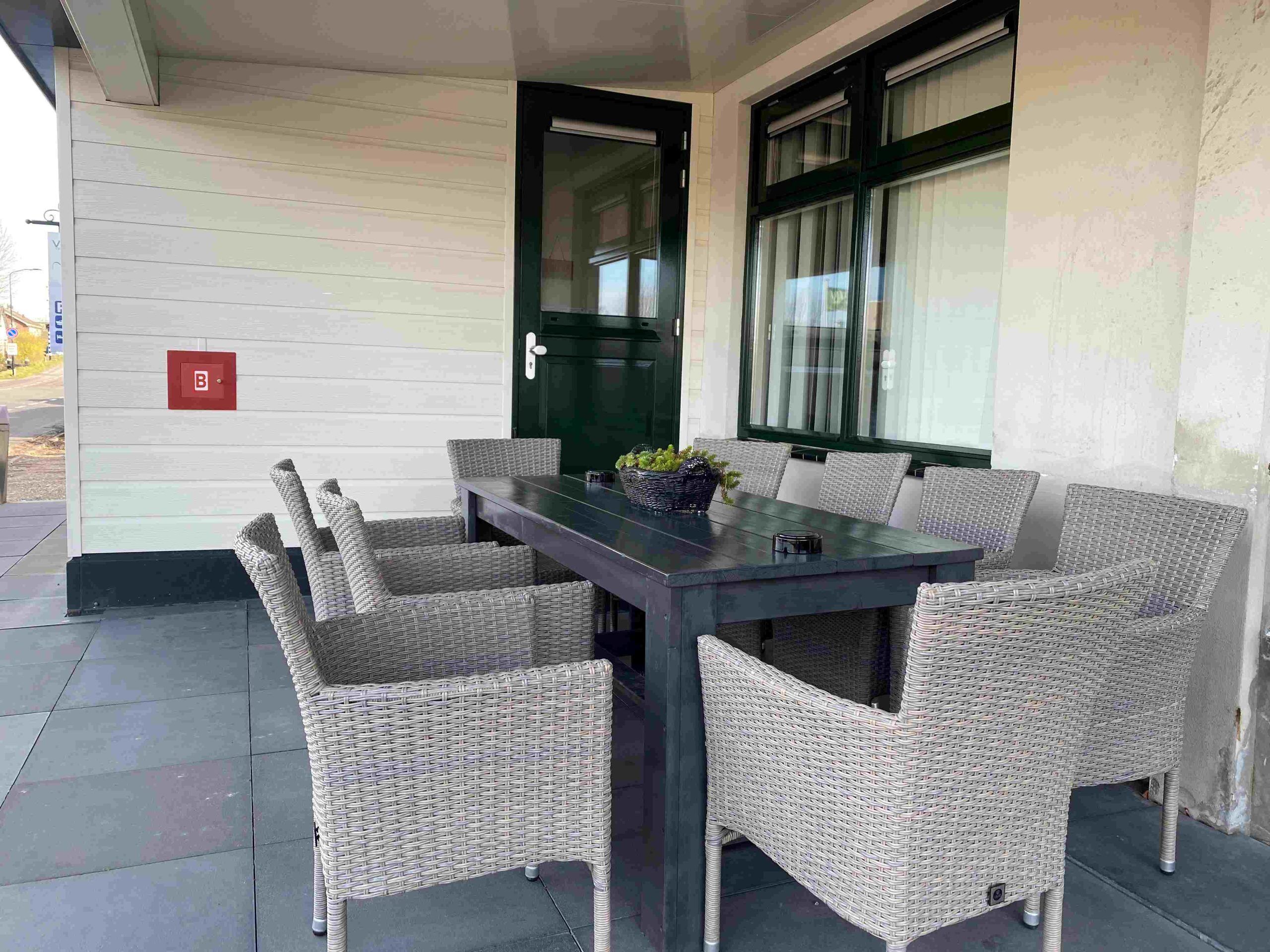 A covered outdoor seating area with a dark table and wicker chairs, a door with a 'B' sign, and large windows.