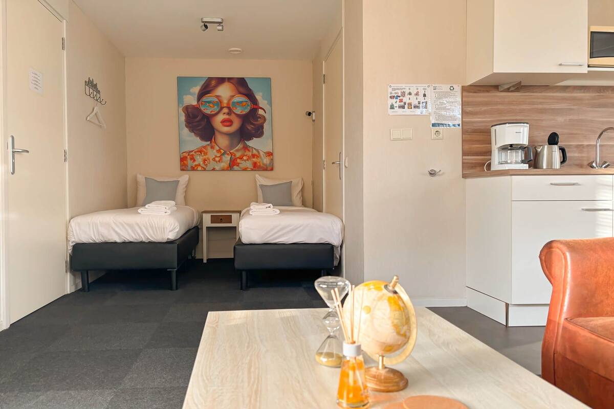 A hotel room with two beds, a small table with a globe and candles, a kitchen area with a coffee maker, and a painting on the wall.