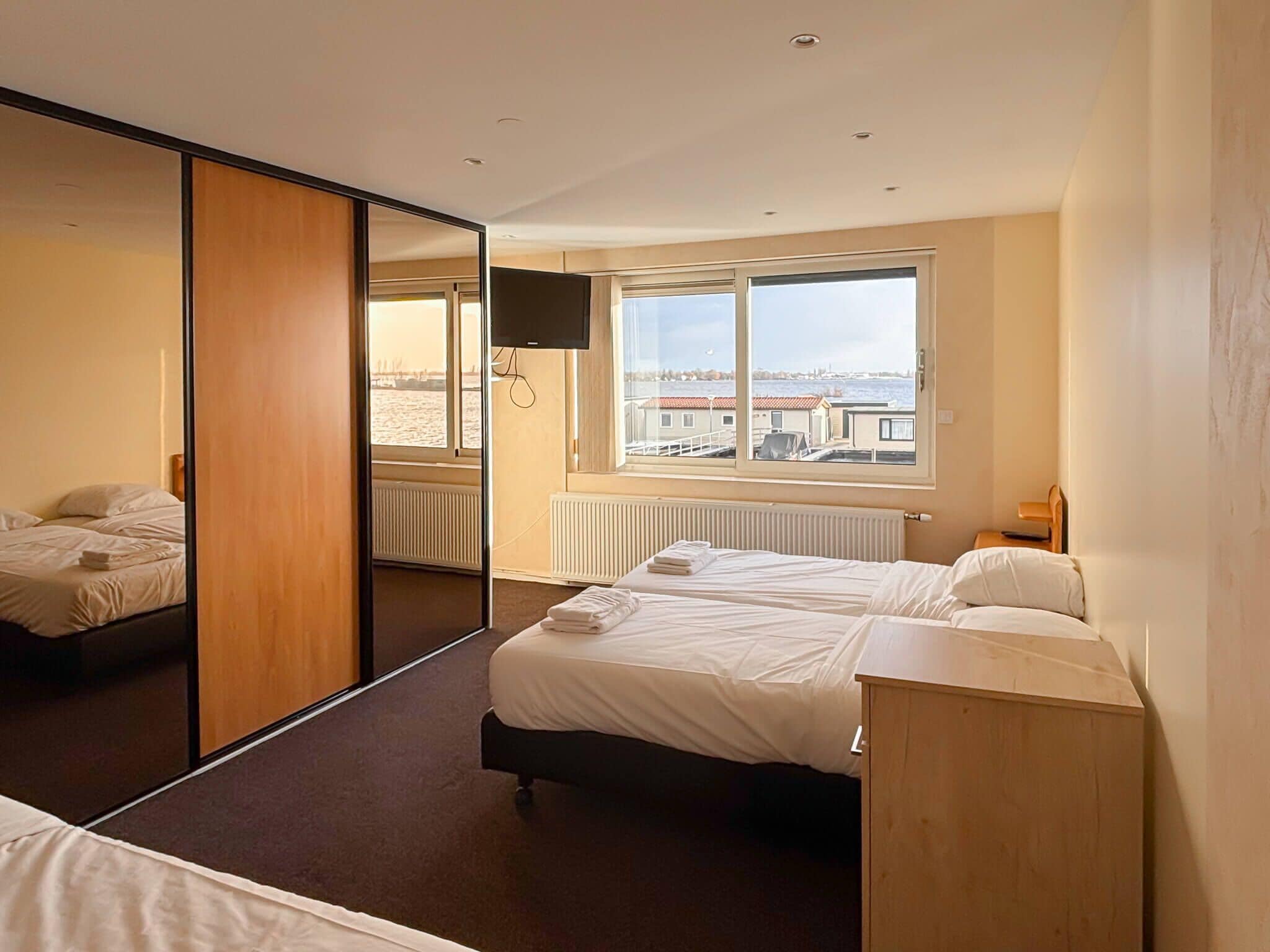 A hotel room with a bed, a wooden wardrobe, a large mirror, a window with a view of the city, and a radiator.