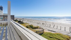 Uitzicht op een strand vanaf een balkon met houten reling, een hek en een heldere lucht.