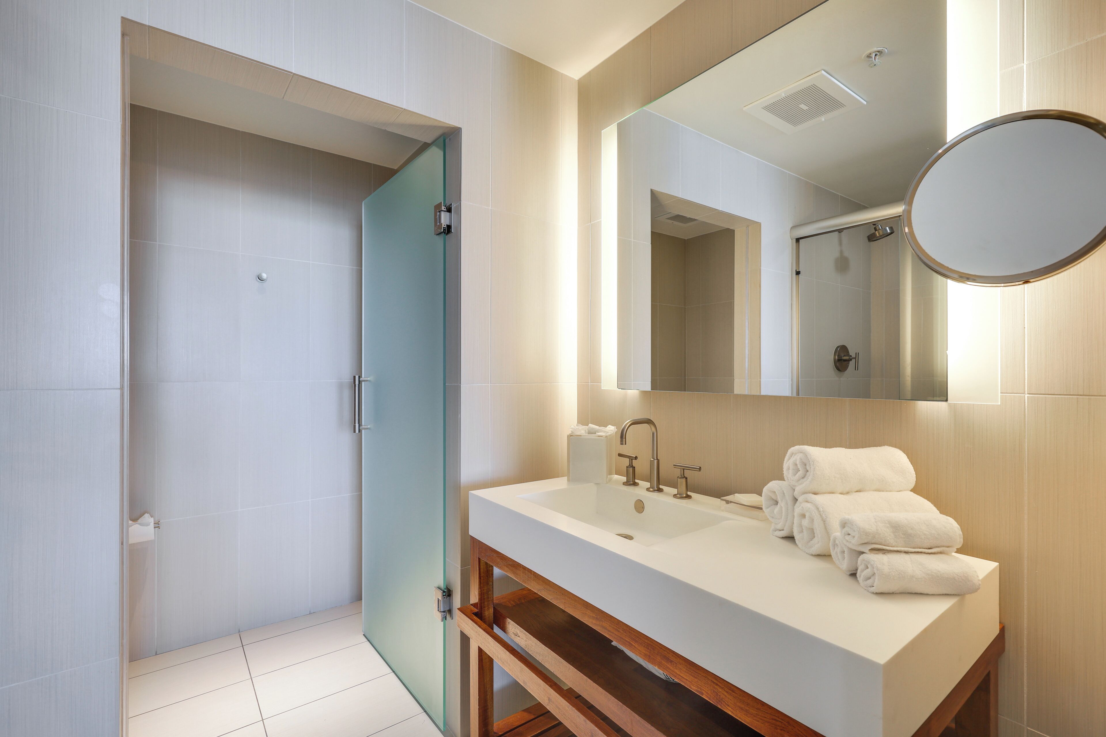 Un baño moderno con mampara de vidrio, un lavabo blanco con tocador de madera y un espejo grande.