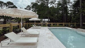 A poolside area with lounge chairs and umbrellas, surrounded by a wooden fence and trees.