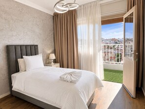 A bedroom with a bed, a nightstand, a lamp, and a view of a cityscape through a balcony.
