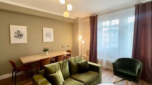A room with a green sofa, a wooden dining table, chairs, a green armchair, and a large window with curtains.