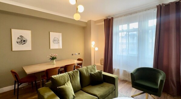A room with a green sofa, a wooden dining table, chairs, a green armchair, and a large window with curtains.