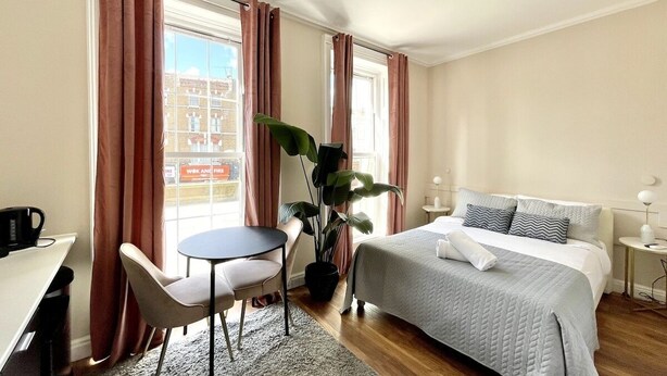 A hotel room with a bed, a desk, a chair, a small table, and a potted plant.