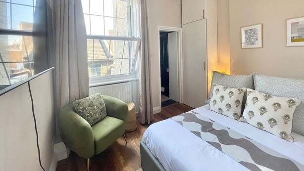 A bedroom with a bed, a green armchair, a window with curtains, and a door leading to another room.