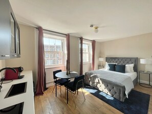 A hotel room with a bed, bedside tables, a desk with a chair, a window with curtains, and a view of buildings outside. - Pretty Studio Flat in Camden (Greater London)