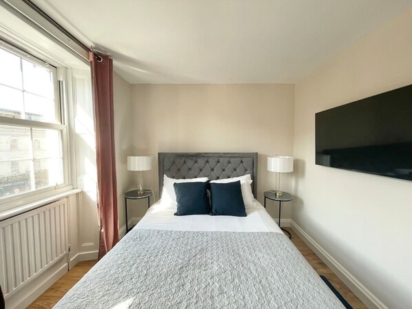 A neatly arranged bedroom with a large bed, two bedside lamps, a television on the wall, and a window with curtains. - Pretty Studio Flat in Camden (Greater London)