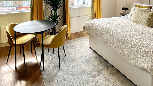 Ett hotellrum med en säng, ett matbord med stolar, en krukväxt och stora fönster med gardiner.