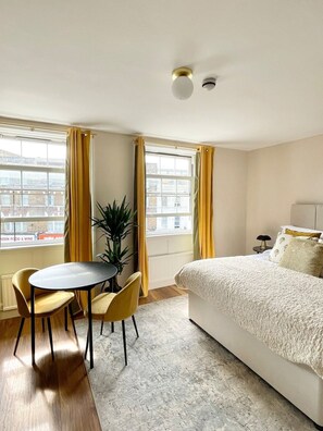 A hotel room with a bed, a dining table with chairs, a potted plant, and large windows with curtains.