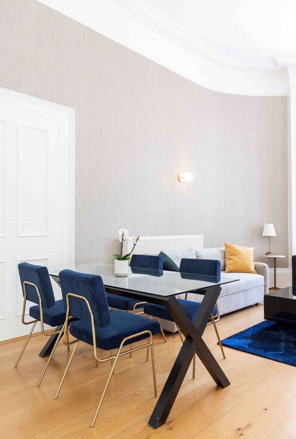 Een moderne woonkamer met een glazen eettafel, blauwe fauteuils, een witte bank met gele kussens en een klein bijzettafeltje met een lamp.
