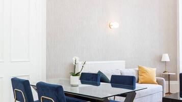 Een moderne woonkamer met een glazen eettafel, blauwe fauteuils, een witte bank met gele kussens en een klein bijzettafeltje met een lamp.