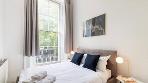A neatly made bed with white linens and two pillows, a bedside table with a lamp, and a window with curtains. - Classy 2 bed apartment with high ceilings (Greater London)