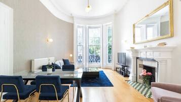 Een ruime woonkamer met een open haard, een eethoek met een glazen tafel en een groot raam met uitzicht op groen.