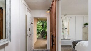 Ein Flur, der in ein Schlafzimmer führt, mit einem Bett, einer Tür, einem Fenster mit Vorhängen und Blick ins Grüne.