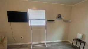 Een kamer met een aan de muur bevestigde televisie, een raam met zonwering, een tafeltje met een lamp en een stoel.