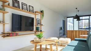 Una sala de estar moderna con un televisor de pantalla plana grande, estanterías de madera, una mesa redonda de madera, un sofá azul y una planta colgante.