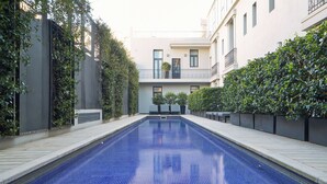 Una piscina con superficie azul de azulejos, rodeada por un seto bajo y un edificio con balcones.
