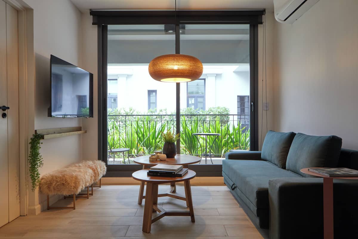 A modern living room with a sofa, a coffee table, a TV mounted on the wall, and a view of a balcony with plants.