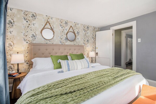 A neatly made bed with a green blanket, a wooden nightstand with a lamp, and a floral wallpaper.