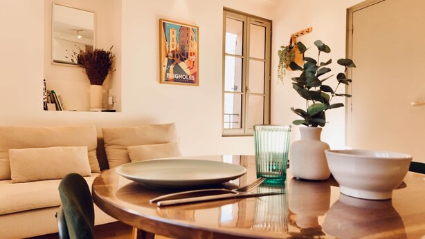 A dining area with a round table, a plate, a bowl, a vase with a plant, and a painting on the wall. - Dubonnet - Coeur de ville (Brignoles)