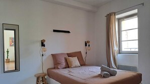 A bedroom with a large bed, a small stool, a mirror, and a window with curtains. - Dubonnet - Coeur de ville (Brignoles)