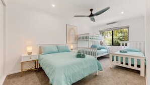 Une chambre avec un lit superposé, un lit simple, une table de chevet, un ventilateur de plafond et une fenêtre donnant sur un paysage verdoyant.