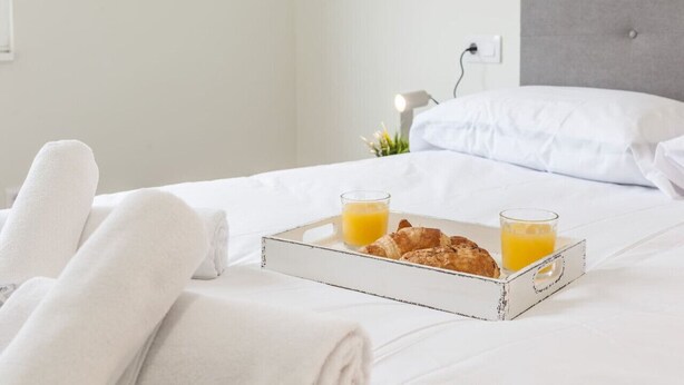 Una cama bien hecha con sábanas blancas, una bandeja con cruasanes y jugo de naranja, y una lámpara de noche.