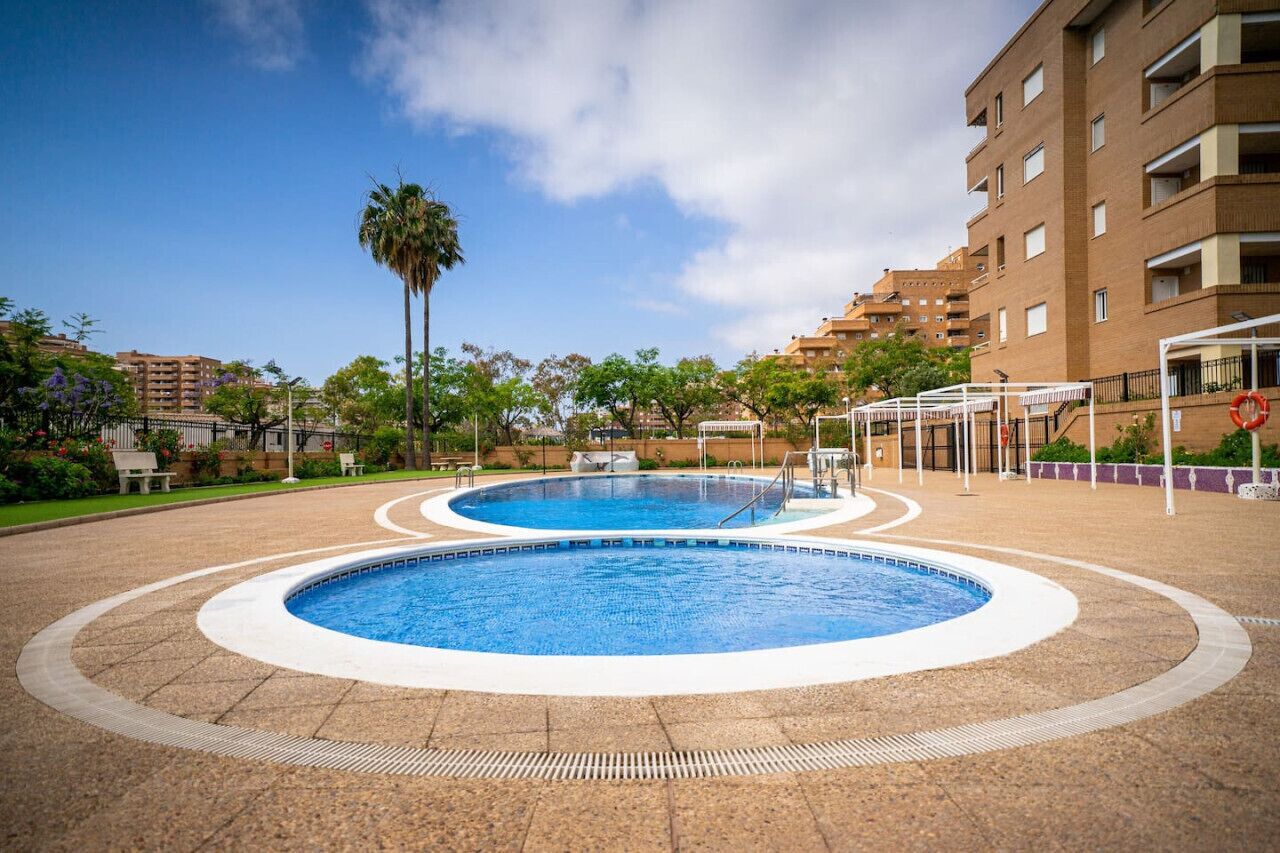 A swimming pool with a circular design, surrounded by a paved area with a palm tree and a building in the background.