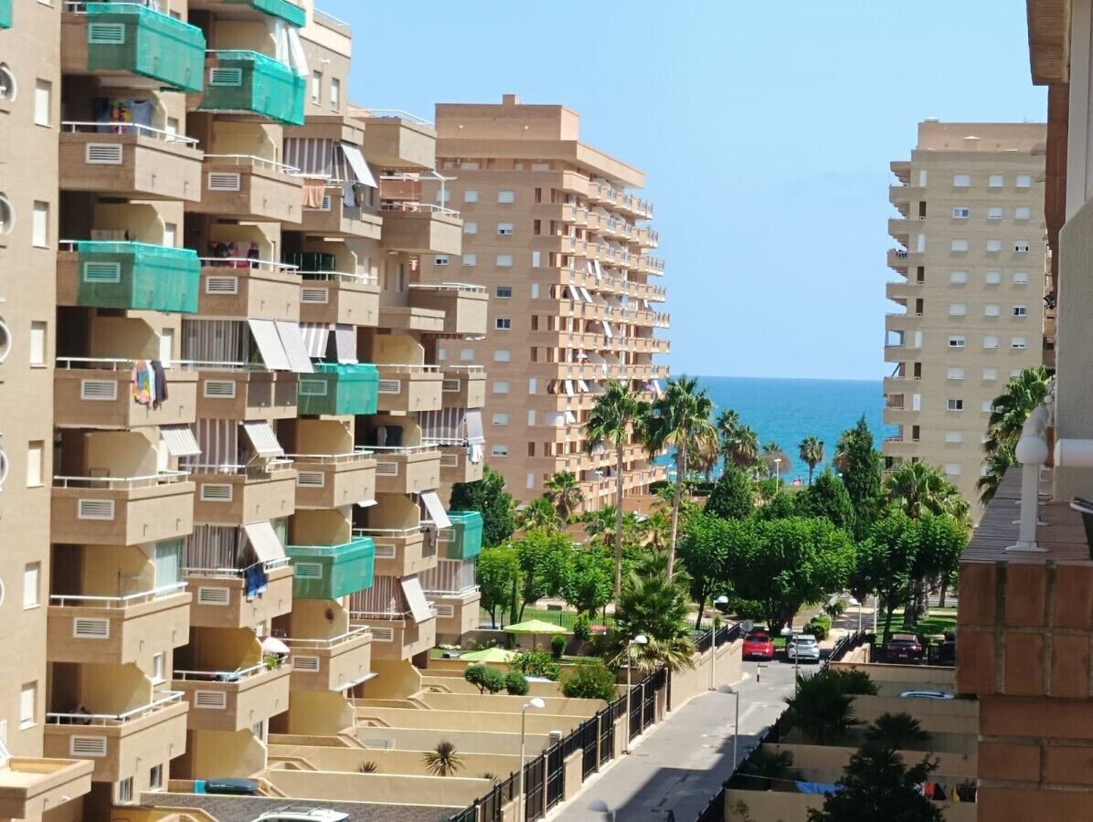 Een hoog appartementencomplex met meerdere balkons, een helder uitzicht op zee, en een verzorgde stadstraat met geparkeerde auto's en groen.