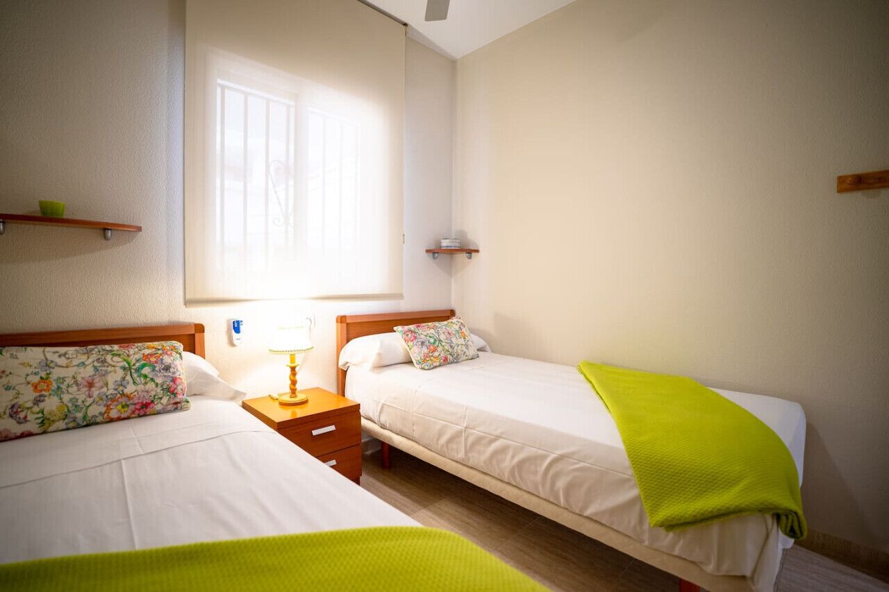Habitación de hotel con dos camas individuales, una mesita de noche con lámpara, una ventana con persianas y un estante pequeño con un cuenco.