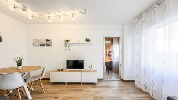A modern living room with a wooden dining table, white sofa, and a TV on a white cabinet. There are plants on the shelves and a staircase leading to another room. - Family-Friendly Stay near Madrid (San Sebastián de los Reyes)