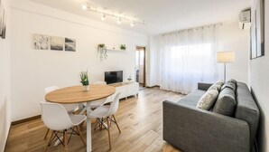 A modern living room with a grey sofa, a wooden dining table with white chairs, a flat-screen TV, and a small plant on the wall. - Family-Friendly Stay near Madrid (San Sebastián de los Reyes)