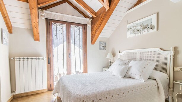 A bedroom with a large bed, wooden beams, a radiator, and a window with curtains. - Rustic Duplex in the Pyrenees - Garden & Parking (Senegüé)