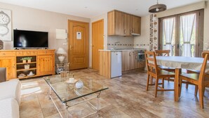 A compact living space with a glass coffee table, wooden dining table, and a kitchen area with wooden cabinets and a built-in oven. - Rustic Duplex in the Pyrenees - Garden & Parking (Senegüé)